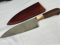 Full view of vintage 7.5-inch chef's knife with wooden handle and leather sheath beside it, showing condition and design.