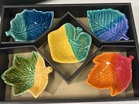 Five small leaf shaped dishes in a black form fitting box, showing a set of different colored glazed ceramic dishes in blue, turquoise, yellow-green, green-yellow, and orange-red, each about 3.5 inches in length with veined leaf details.