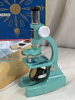 Close-up angle of vintage Tasco child’s microscope showing turquoise body, white focus knobs, black eyepiece, and stage with clips.