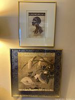View of two framed art prints hung on wall, one above the other: the upper is a portrait etching of an Ethiopian woman, the lower is a decorative print of quails and flowers with a patterned border and gold frame.