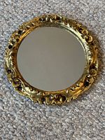Small golden ornate wooden framed circular mirror on carpet showing carved details and reflective surface.