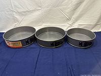 Photo of three round springform pans of varying sizes laid out on a blue cloth background, showing front clasps and removable bottoms.