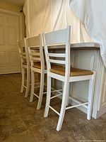 Three white wood bar or island chairs with woven wicker seats placed against a kitchen counter.