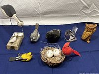 All seven bird figurines displayed on a blue cloth, showing variety of styles and mediums, including wood, pottery, glass, and ceramic.