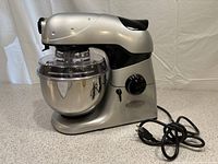 Side view of silver and black stand mixer with attached stainless steel mixing bowl and clear plastic lid