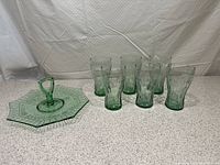 Set of a green depression glass serving plate with octagonal shape and keyhole handle to the left, and six green-tinted Coca-Cola glasses to the right, all placed on speckled surface.