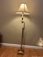 Full view of the brass floor lamp standing against a wall, lit and showing the square base and segmented pole design.