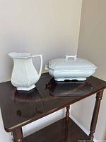 Photo of the white ironstone pitcher with ribbed lower half, flared spout, and handle alongside the rectangular Royal Ironstone tureen with lid and decorative handle, on a dark wood table.