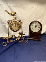 Photo showing both clocks: the gold metal ornate table clock with birds and floral design on the left and the wooden Bombay Company mantle clock on the right.