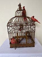 Front view of brown metal bird cage with three red artificial birds perched on and outside the cage.