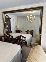 Large rectangular wall mirror hung on a wall reflecting a dining room with a table, chairs, chandelier, and cabinet. Shows overall size and setting of mirror.