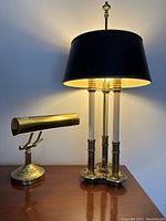 Photo of the gold tone banker's lamp and the 3-arm candlestick table lamp with black shade lit on a wooden surface.