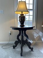 Full view of lamp on vintage oval wooden table showing lamp lit with cream shade and ornate dark wood table base and top