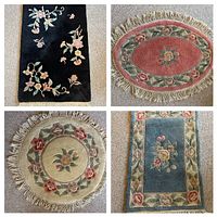 Four wool floral accent rugs arranged on a carpeted floor, showing black, pink, beige, and blue pieces with floral patterns and fringed edges.