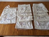 Ten unopened natural-colored canvas drawstring bags with embroidered dress mannequin designs and 'Arianne' branding, stacked and laid out on a wooden surface.