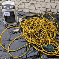 Portable MaxHeat electric heater, long yellow extension cord, and white outlet extension box laid on a black plastic mat on a stone surface.