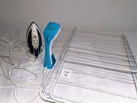 Three laundry items on a white surface: white drying rack, blue and white Black & Decker handheld steamer, and white and black T-fal iron, cords visible.