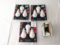 Three unopened 3-packs of FEIT Electric 60W dimmable warm white LED bulbs plus one opened box of Sylvania daylight CFL bulbs with 2 bulbs remaining shown in packaging