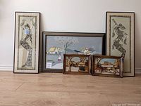 Five framed art pieces against a plain wall including two Balinese tapestries, two Syroco 3D winter ride scenes, and one farm scene in frames.