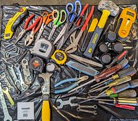 Top-down photo of assorted hand tools including hammers, scissors, screwdrivers, tape measures, pliers, wrenches, hex keys, clamps, and a utility knife arranged on a black background.