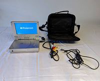 Open Polaroid portable DVD player with built-in screen displaying Polaroid logo, showing control buttons, with black carrying case and cables on white fabric background.