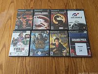 Photo showing all eight original PS2 game cases arranged on wooden floor: Mortal Kombat Shaolin Monks, Mortal Kombat Armageddon, Mortal Kombat Deception, Gran Turismo 4, FIFA 06, Lord of the Rings The Two Towers, Harry Potter and the Chamber of Secrets, Grand Prix Challenge.