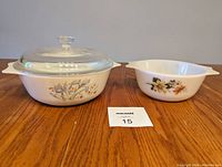Photo showing two white Pyrex bowls with floral designs side by side on wooden table. Larger one has a glass lid.
