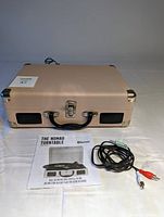 Closed beige portable suitcase record player with black handle, metal corners, front lock, and built-in speakers. Manual and cables placed in front.