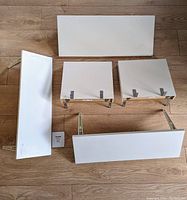 Top view of 5 white shelves arranged on floor showing size and bracket placement, including longer BURHULT and Canadian Tire shelves and smaller LACK shelves.