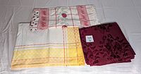 Three vintage rectangular table covers laid flat on a white surface. Left cover is yellow and white checkered with orange stripes, top cover is white with red stripes and strawberry design, and right cover is burgundy with floral damask pattern.