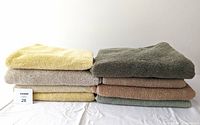 Stack of 8 cotton towels in yellow, light brown, and green colors positioned side by side on a white surface.