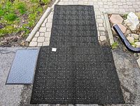 Two large black carpet mats with patterned textured surface and one smaller blue door mat placed outdoors on stone tiles.
