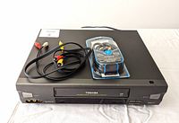 Black Toshiba VHS VCR with front VHS slot, control buttons and RCA ports, accompanied by RCA and coaxial cables on top of VCR.