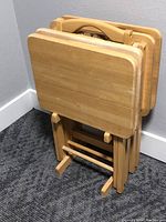 Four solid wood TV trays folded and stored upright on matching wooden stand on a carpeted floor against a wall.