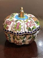 Vintage cloisonné enamel lidded trinket box on brown surface showing floral enamel pattern and gold-tone finial on lid.