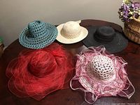 Five vintage summer hats displayed on a table including teal woven, off-white cut-out, black lace-like, red mesh with feathers, and white with pink ruffles hats.