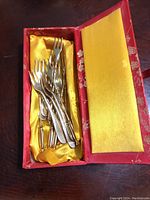 Set of ten gold plated hors d’oeuvre forks placed inside red silk box lined with yellow satin fabric.
