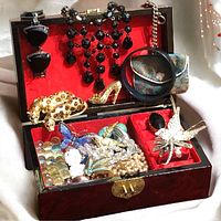 Open wooden jewelry box showing a mix of costume jewelry including brooches shaped like leopard and butterfly, beaded necklaces, and a small watch.