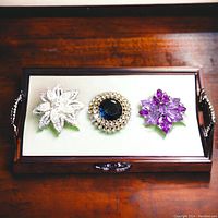 Three vintage rhinestone brooches displayed in an open wooden box with white lining: a clear rhinestone flower, a round blue and clear rhinestone brooch, and a purple rhinestone flower.