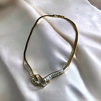 Full view of necklace lying on white satin fabric, showing gold-tone snake chain with central wavy design set with clear baguette stones.