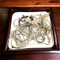 Top view of multiple silver chains and bracelets displayed on a white square plate with wooden frame showing mixed designs and decorative elements