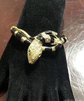 Photo of vintage snake bracelet featuring gold-tone metal with black enamel stripes and crystal detailing on the snake's head with green rhinestone eyes. Displayed on black fabric.