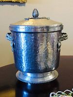 Full view of vintage silver-plated ice bucket with hammered texture, sculpted handles, lid with decorative finial, and rounded base.