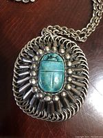 Close-up front view of oval silver tone pendant with turquoise scarab centerpiece and decorative metal surround on linked chain.