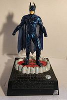 Front view of a Batman figurine piggy bank with a metallic blue finish, standing on a detailed base with a brick pattern and red and white accents. Visible bent ear and missing battery cover on the base