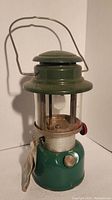 Green Coleman Model 335 lantern standing upright, showing wire bail handle, glass globe, red control knob, and metal base with some dirt and rust spots