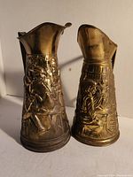 Both copper jugs side by side showing embossed detailed scenes of figures and objects on their exteriors.