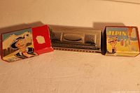 Front view of the metal Alpina Koch harmonica placed between two halves of the original box with colorful alpine-themed illustration, showing the harmonica and box condition.