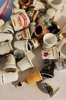 Photo shows a pile of assorted thimbles including ceramic souvenir pieces with painted place names and floral designs, a copper colored metal thimble and a wooden thimble with handle.