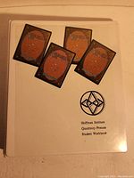 Photo of closed white 3-ring binder labeled Hoffman Institute Quadrinity Process Student Workbook with some Magic The Gathering cards placed on top, showing backs of the cards.
